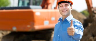 un homme avec une chemise bleue et un casque de chantier lève le pouce en l'air, sourire aux lèvres. Derrière lui, un engin de chantier.