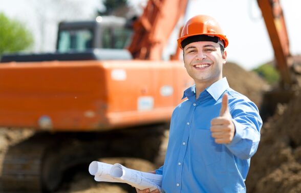 un homme avec une chemise bleue et un casque de chantier lève le pouce en l'air, sourire aux lèvres. Derrière lui, un engin de chantier. - Agrandir l'image, fenêtre modale