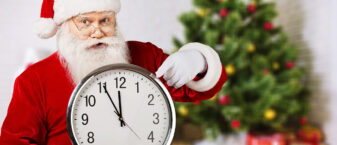 un père noël montre une horloge, un sapin et des cadeaux derrière lui.